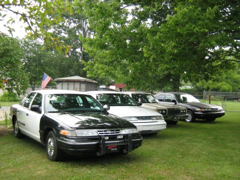 1988 Mercury Grand Marquis - Four sedans, including a police car, are parked on a lawn under trees.