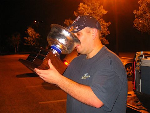 1985 Ford LTD Crown Victoria - A man celebrates with a trophy, with a car parked nearby at night.