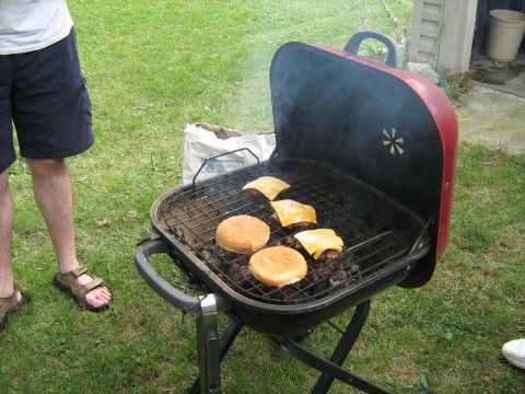 1988 Mercury Grand Marquis - Burgers and cheese melt on a grill as a person stands by, with smoke rising from the coals.