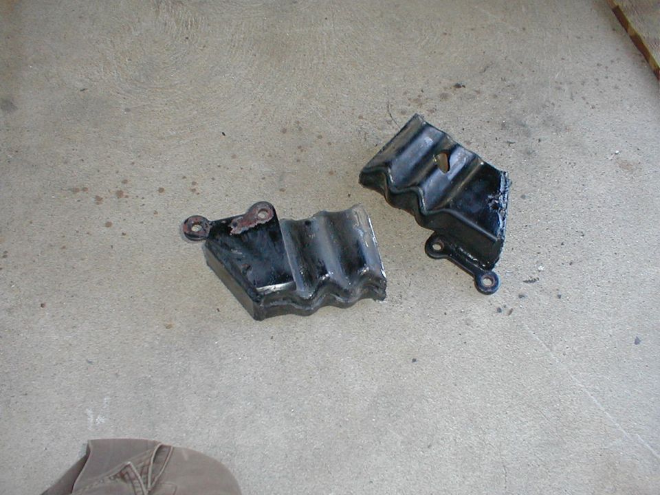 Two broken black metal parts with mounting holes on a concrete surface