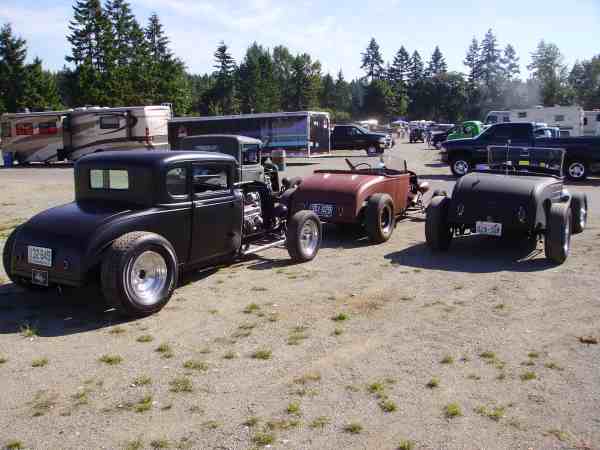 Flat black chopped coupe beside brown and black fenderless roadsters on a gravel lot