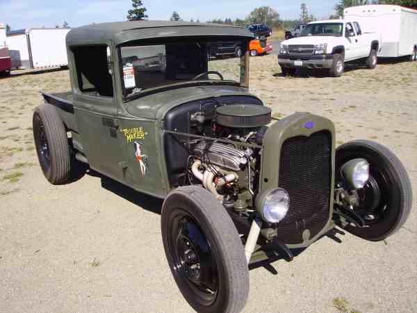 Olive green hot rod pickup truck with exposed engine and wide rear tires. Text on door reads 'Trouble Maker'