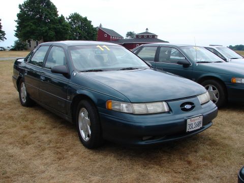 1993 Ford Taurus - A 1993 Ford Taurus is parked on dry grass with other vehicles and barns in the background.