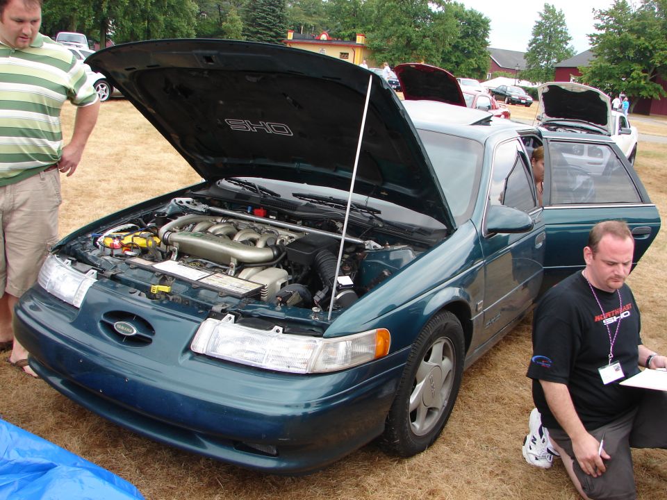 Green 1993 Ford Taurus SHO with hood open, showing engine. Man in black shirt kneels nearby