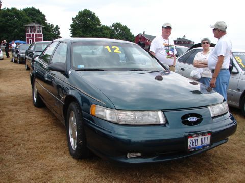 1993 Ford Taurus - A dark green Ford Taurus is parked on dry grass with people standing nearby at an outdoor event.