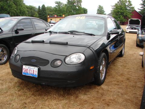 1993 Ford Taurus - A black Ford Taurus with a car cover and a '96 sticker is parked on grass next to other vehicles.