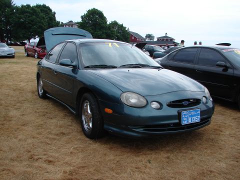 1993 Ford Taurus - A teal sedan with its hood open is parked on dry grass at an outdoor event.