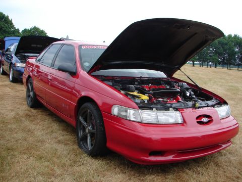 1993 Ford Taurus - A red 1993 Ford Taurus is displayed with its hood open, revealing the engine bay, alongside other vehicles.