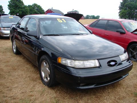 1993 Ford Taurus - A black sedan is parked on dry grass, with other vehicles nearby and barns in the background.