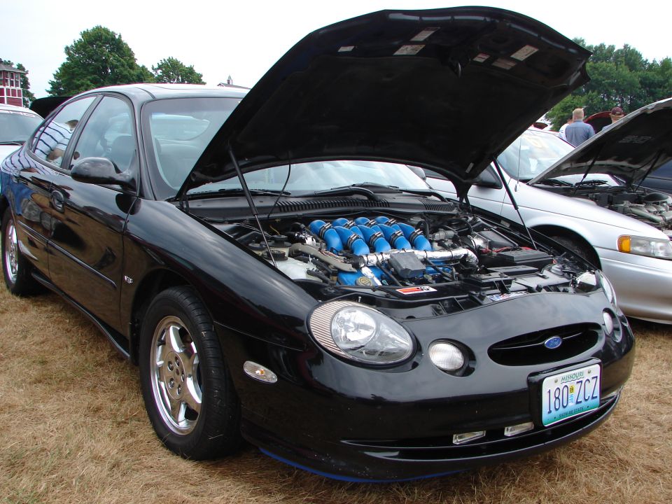 Black Ford Taurus with hood open, showing a modified engine with blue intake runners. License plate 180 ZCZ