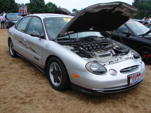 1993 Ford Taurus - This sedan has a V8 engine visible under its open hood, parked on dry grass at an outdoor event.