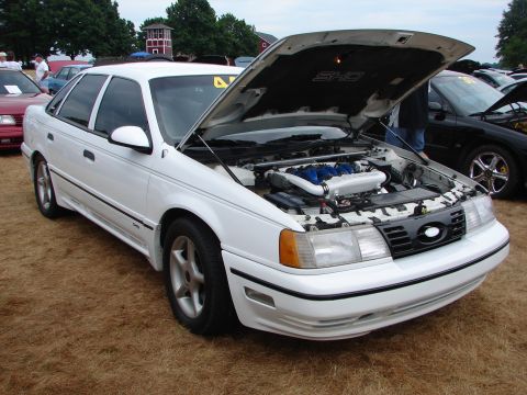 1993 Ford Taurus - A white 1993 Ford Taurus SHO is displayed with its hood open, revealing the engine bay at an outdoor car show.