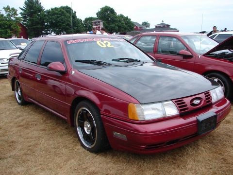 1993 Ford Taurus - A maroon sedan with a dark hood and custom rims sits on dry grass at an outdoor event.