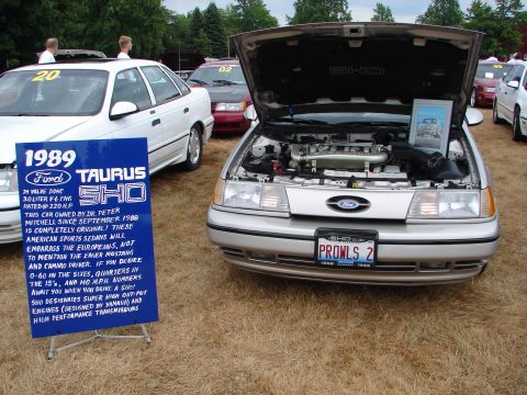 1993 Ford Taurus - A silver 1989 Ford Taurus SHO is displayed with its hood open, alongside another Taurus and a descriptive sign.