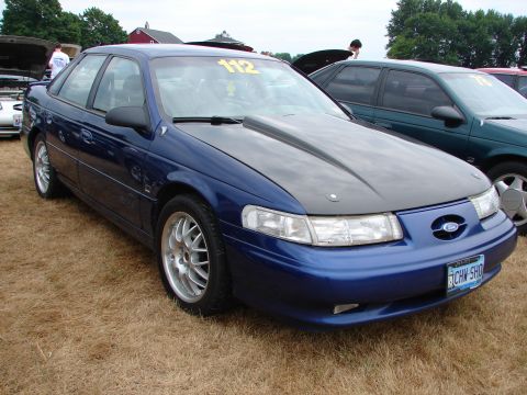 1993 Ford Taurus - A blue 1993 Ford Taurus is shown with a custom hood and racing numbers, parked at an outdoor event.