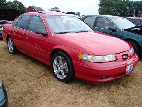 1993 Ford Taurus - Red sedan with a '92 sticker on the windshield, parked on grass at an outdoor event.