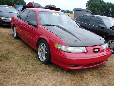 1993 Ford Taurus - A red sedan with a black hood and racing number 49 is parked on grass next to other vehicles.