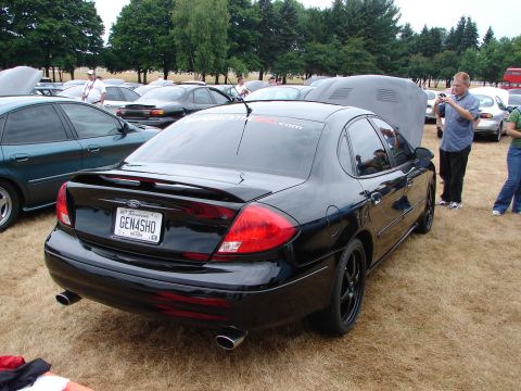 1993 Ford Taurus - A black sedan is parked on dry grass with other cars and people around.