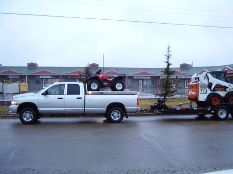 2005 Dodge 3500 - A silver 2005 Dodge 3500 pickup truck is seen from the side, towing a trailer carrying a skid steer loader and an ATV.