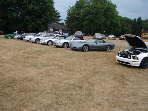 1993 Ford Taurus - A variety of cars are parked on a dry, grassy field with trees and a building in the background.