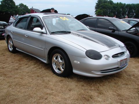 1993 Ford Taurus - A silver sedan with racing stripes is parked on dry grass at an outdoor car show.