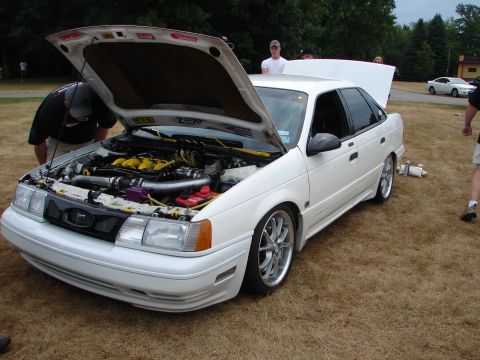 1993 Ford Taurus - A white sedan with its hood open reveals a modified engine, custom wheels, and lowered suspension.
