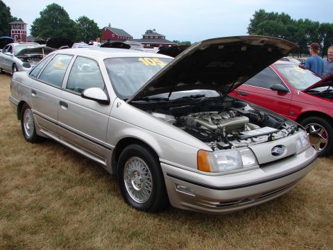 1993 Ford Taurus - A silver sedan with its hood open is parked on a grassy field next to other vehicles.