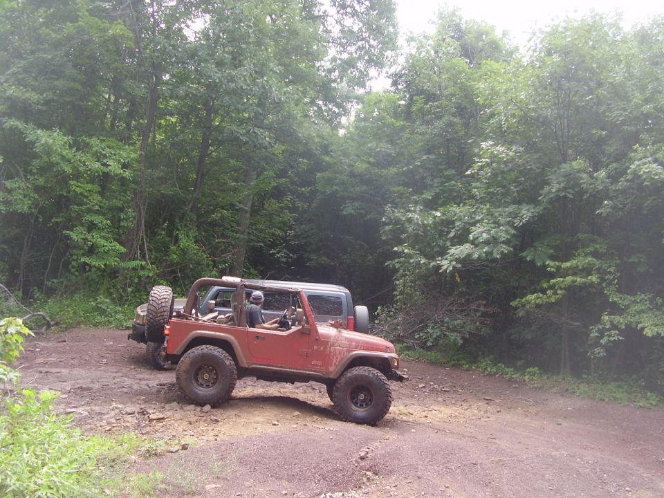 Topless red Jeep Wrangler on mud tires parked on a dirt trail in green woods