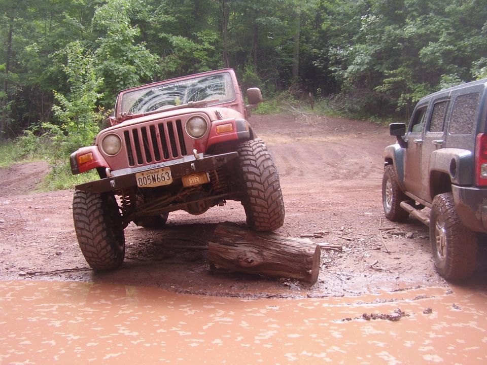 2003 Jeep Wrangler with lifted suspension driving over a log in a muddy puddle, another SUV is parked nearby