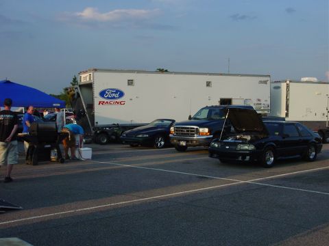 1986 Ford Mustang - A black 1986 Ford Mustang hatchback is parked with its hood open, alongside a Ford Racing trailer and other vehicles.