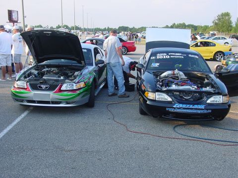 1986 Ford Mustang - Two Ford Mustangs are parked side-by-side at a drag strip with their hoods open, revealing their engines.