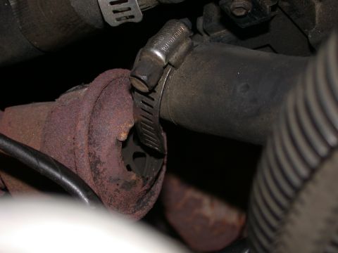 1987 Lincoln Town Car - A close-up view of a rusty exhaust pipe connection, held in place by a metal hose clamp on a dark rubber hose.
