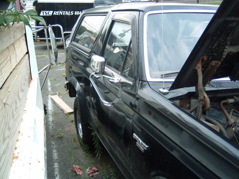 1986 Ford Bronco - A black Ford Bronco is parked next to a wooden fence, with its hood open revealing the engine bay.