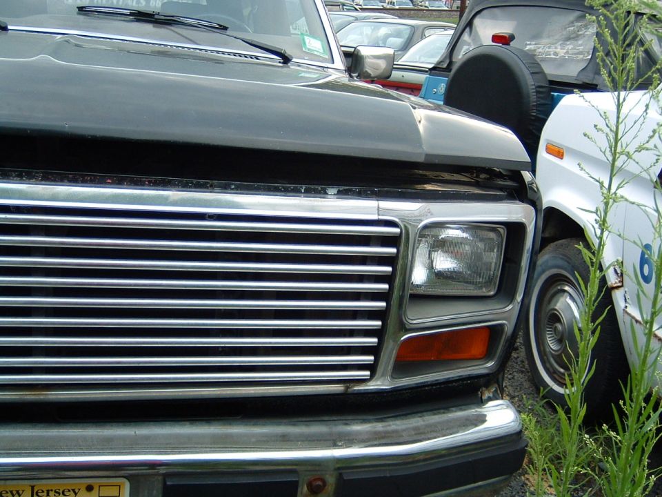 Front grille and headlight of a dark-colored Ford truck, parked next to a white vehicle