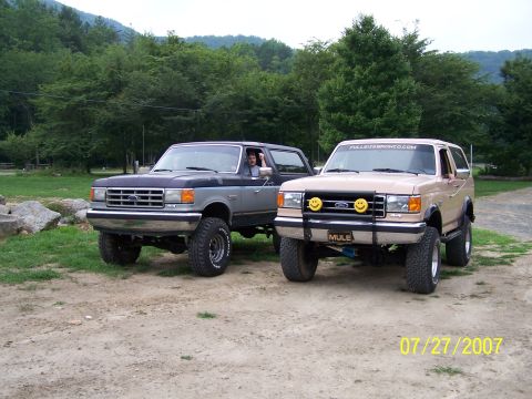 1979 Ford F-150 - Two Ford Broncos are parked outdoors on a dirt road with a wooded hillside behind them.
