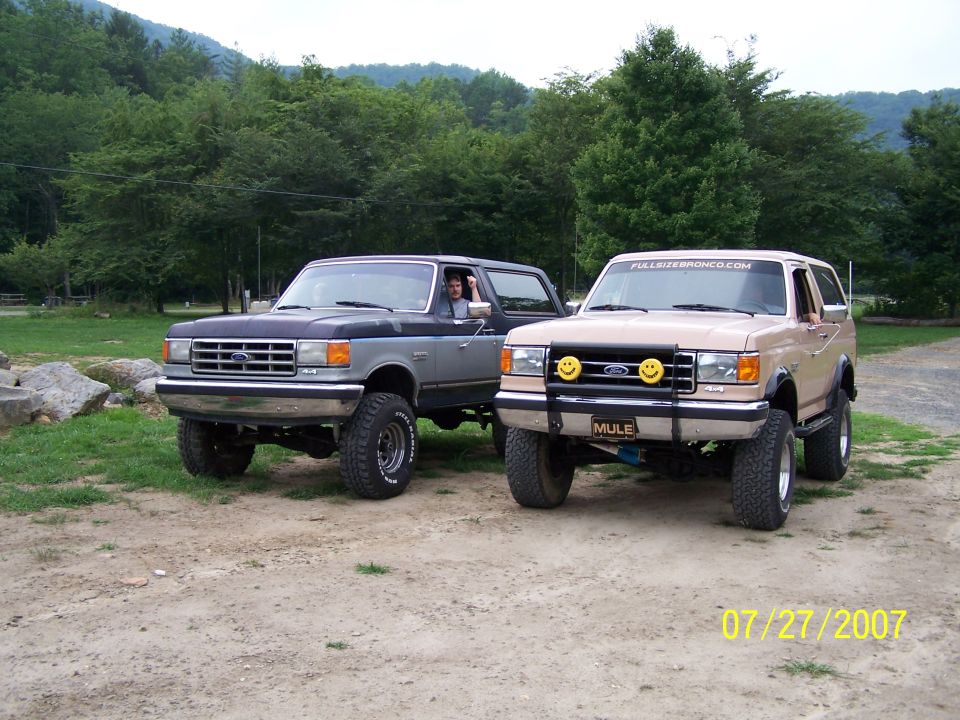 Two lifted Ford Broncos, one tan and one blue/silver, parked on a dirt road with trees and hills in the background. The tan Bronco has "MULE" on the license plate and "FULLSIZEBRONCO.COM" on the windshield banner