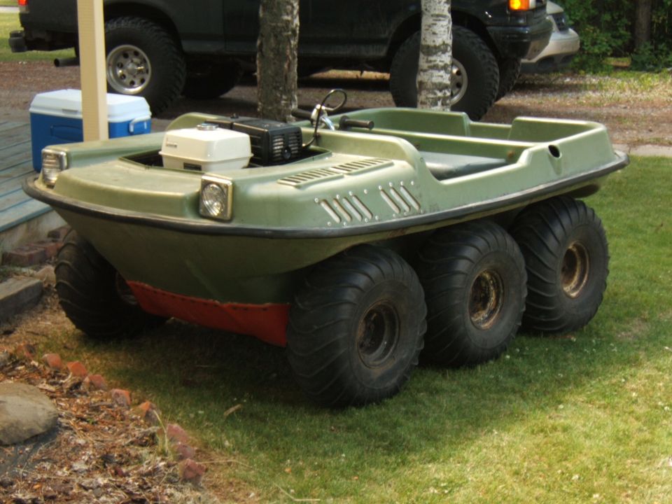 Green 6-wheel amphibious ATV with red hull on grass, front 3/4 view