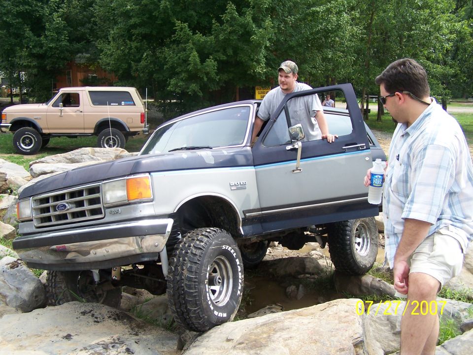 Two Ford Broncos off-roading over rocks. One is a tan Bronco in the background, and the other is a two-tone gray and blue Bronco in the foreground with a man leaning out the door