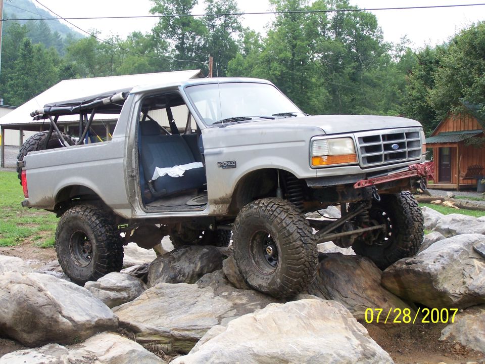 Modified Ford Bronco with off-road tires navigating large rocks. The driver's door is open, revealing the interior
