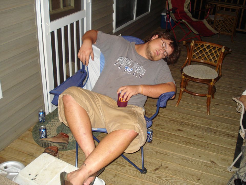 Man sleeping in a blue folding chair on a wooden deck, holding a red cup