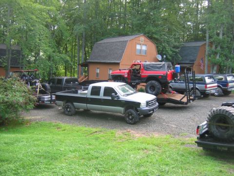 1992 Ford Mustang - A collection of trucks and off-road vehicles are parked in a gravel lot near houses and trees.