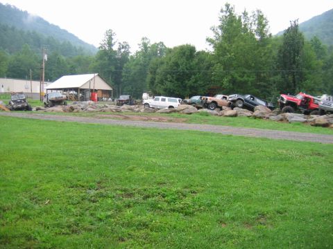 1992 Ford Mustang - A collection of modified off-road vehicles are parked on a rocky terrain, with a building and wooded hills visible behind them.