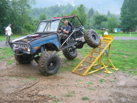 1992 Ford Mustang - A heavily modified blue off-road vehicle attempts to climb a yellow ramp in a muddy outdoor setting.