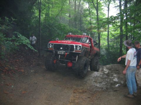 1992 Ford Mustang - A heavily modified red truck navigates a challenging muddy terrain in a wooded area.
