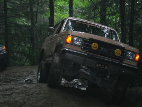 1992 Ford Mustang - A lifted Ford Bronco navigates a muddy off-road trail in a wooded area, with another vehicle visible to the left.