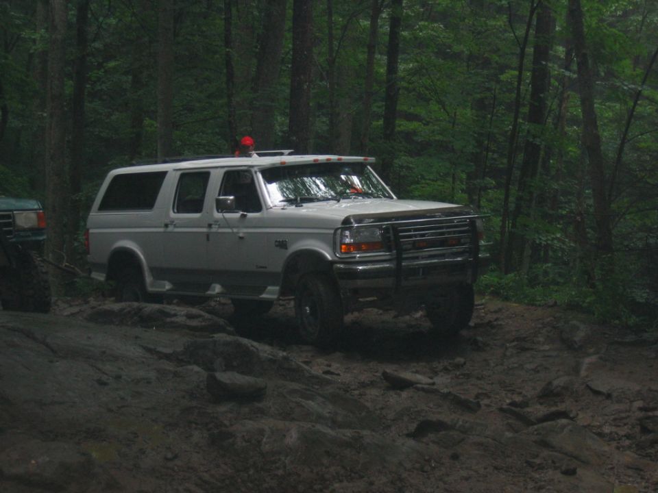 White Ford Bronco SUV on a rocky, muddy trail in the woods. Another green vehicle is partially visible to the left