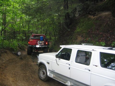 1979 Ford Bronco - Two off-road vehicles navigate a narrow dirt path surrounded by dense forest foliage.