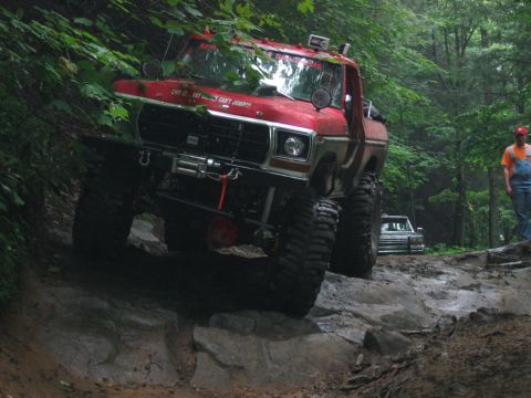 1992 Ford Mustang - Modified red Ford F-Series pickup truck navigates a challenging rocky path in a wooded area, with another vehicle visible in the background.