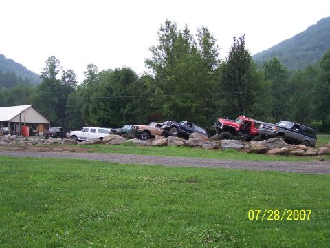 1979 Ford F-150 - Several modified trucks and SUVs are positioned on a rocky terrain, with a building and trees in the background.