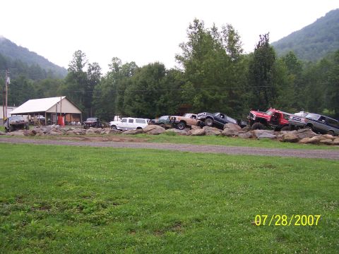 1979 Ford F-150 - Multiple off-road vehicles are positioned on a rocky terrain with a building and trees in the background.